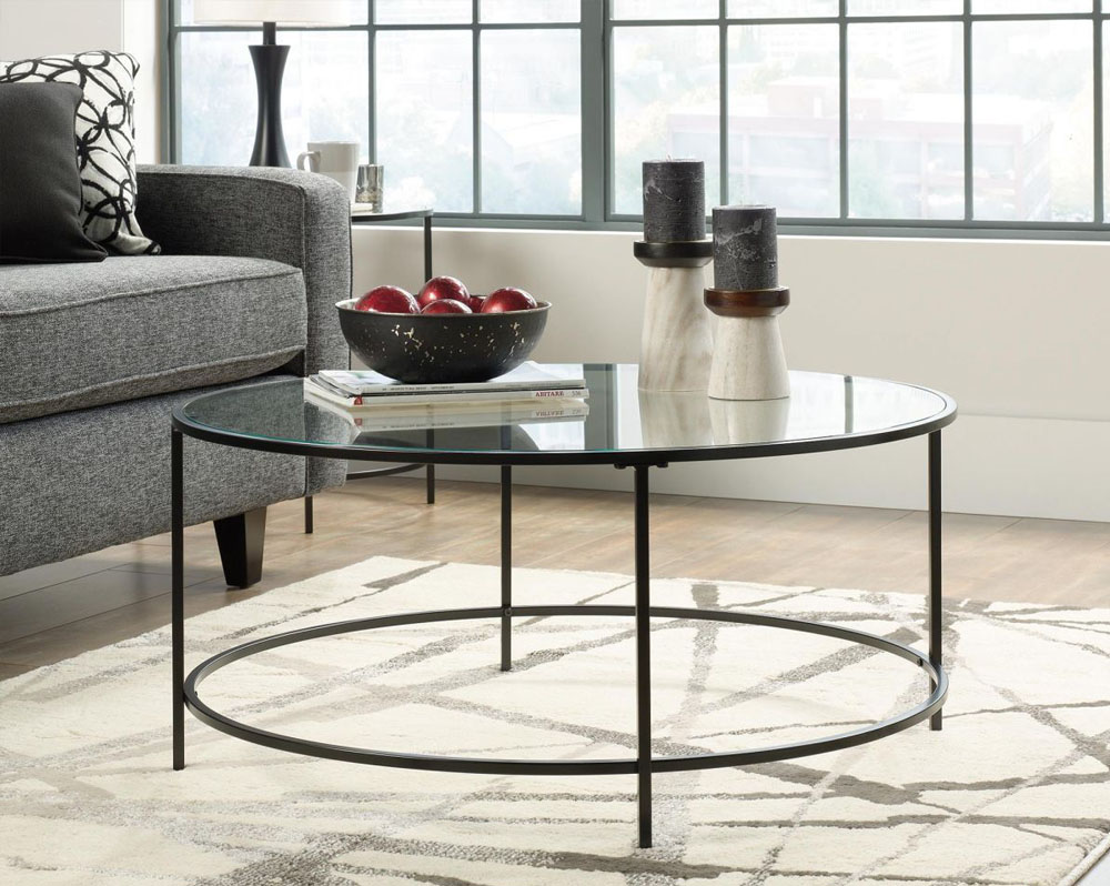 Round glass top coffee table with black frame in front of a grey sofa